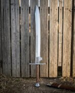 Hand forged bastard sword with a broad engraved double edged blade and disc pommel standing point up against a weathered timber fence beside a leather scabbard