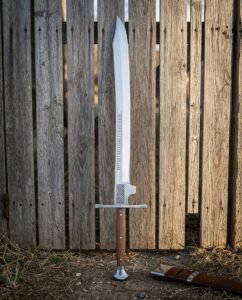 Hand forged bastard sword with a broad engraved double edged blade and disc pommel standing point up against a weathered timber fence beside a leather scabbard