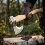 Hand forged bearded axe with etched knotwork head and pale birch haft held in a gloved hand above a birch log and leather sheath