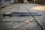 Hand forged black Damascus Viking sword, full length side view on weathered flagstones with its scabbard laid alongside