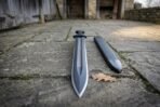 Hand forged black Damascus Viking warrior sword pointing down on stone flagstones in a courtyard beside its black scabbard