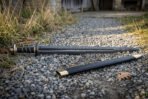 Hand forged blackened high carbon steel Viking sword, low full length view on gravel next to its black scabbard and dry grass