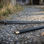 Hand forged blackened high carbon steel Viking sword, low full length view on gravel next to its black scabbard and dry grass