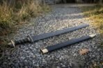 Hand forged blackened high carbon steel Viking warrior sword lying on a gravel path beside its black scabbard with brass tip