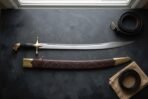 Hand forged curved sword with a broad polished scimitar blade, brass cross guard and leather grip, laid on dark slate beside its tooled leather scabbard and belts