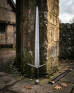 Hand forged damascus arming sword with an etched rune inscription and quatrefoil cross guard standing upright against a weathered sandstone wall beside its leather scabbard