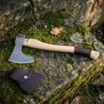 Hand forged damascus bearded axe with a layered etched steel head and pale birch haft, laid on a mossy stone step next to its stitched leather head sheath