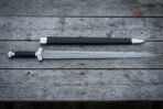 Hand forged Damascus Chinese sword with a straight layered double edged jian blade and wrapped grip lying on weathered timber decking beside a black scabbard