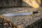 Hand forged Damascus cutlass with a broad layered blade, open brass knuckle bow and ribbed black leather grip resting on a weathered limestone ledge in front of an old stone wall
