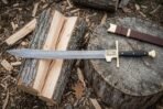Hand forged Damascus falchion with a layered patterned blade and gilt brass ring guard resting on a split log and tree stump beside a brown leather scabbard