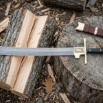 Hand forged Damascus falchion with a layered patterned blade and gilt brass ring guard resting on a split log and tree stump beside a brown leather scabbard