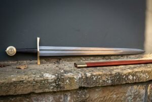Hand forged Damascus knightly sword with a broad forge welded blade and arched brass cross, resting with its russet scabbard on a lichened sandstone ledge.