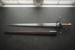 Hand forged damascus rapier with a layered wood grain blade and blackened swept hilt with brass finials laid on dark slate beside its oxblood leather scabbard