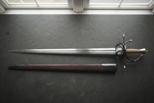 Hand forged damascus rapier with a layered wood grain blade and blackened swept hilt with brass finials laid on dark slate beside its oxblood leather scabbard