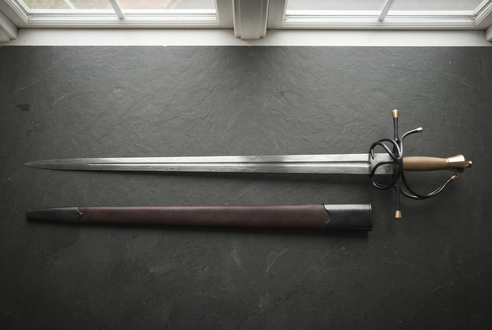 Hand forged damascus rapier with a layered wood grain blade and blackened swept hilt with brass finials laid on dark sla Hand forged damascus rapier with a layered wood grain blade and blackened swept hilt with brass finials laid on dark slate beside its oxblood leather scabbard