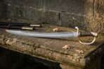 Hand forged Damascus sabre with a layered curved blade, ornate three bar brass hilt and brass wired leather grip lying on a mossy stone garden bench beside its black leather scabbard