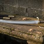 Hand forged Damascus sabre with a layered curved blade, ornate three bar brass hilt and brass wired leather grip lying on a mossy stone garden bench beside its black leather scabbard