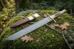 Hand forged Damascus seax blade shown point first across green moss, with the antler handle and leather sheath resting behind