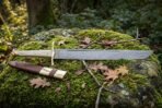Hand forged Damascus seax with a layered single edged blade and pale polished bone handle lying on a moss covered boulder beside its brown leather sheath