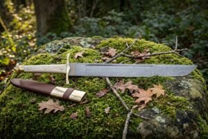 Hand forged Damascus seax with a layered single edged blade and pale polished bone handle lying on a moss covered boulder beside its brown leather sheath