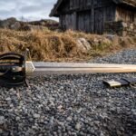 Hand forged damascus short sword with a wood grain layered blade and blackened swept hilt resting on a gravel path beside its black leather scabbard