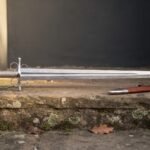 Hand forged damascus side sword with a broad layered leaf shaped blade and polished four ring hilt on a sandstone step beside its russet leather scabbard