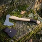 Hand forged Damascus steel Viking bearded axe with leather wrapped grip, lying on a mossy stone step beside its brown leather sheath