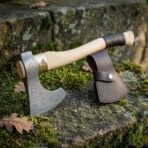 Hand forged Damascus steel Viking bearded axe with patterned head, standing upright on mossy stone next to its brown leather sheath
