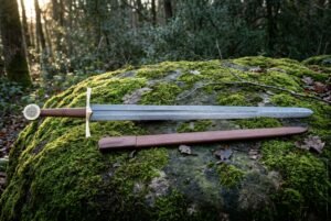 Hand forged Damascus sword with scabbard, broad forge welded blade and carved bone Vegvisir pommel, laid across a moss covered woodland boulder at golden hour.
