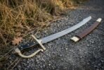 Hand forged Damascus szabla with a layered curved blade, rune cut brass knuckle bow and black horn grip lying on a gravel path beside dry grass and its brown leather scabbard