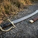 Hand forged Damascus szabla with a layered curved blade, rune cut brass knuckle bow and black horn grip lying on a gravel path beside dry grass and its brown leather scabbard