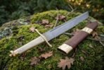 Hand forged Damascus Viking seax with brass fittings, full length view on moss with fallen leaves beside its brass tipped leather scabbard