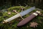 Hand forged Damascus Viking seax with pale grip and brass pommel, close view of the handle end on moss by the scabbard