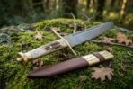Hand forged Damascus Viking seax with stag antler handle and brass guard, lying on moss beside its brown leather sheath in woodland