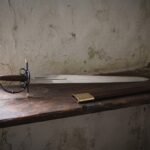 Hand forged espada ropera with a broad polished blade and blackened brass capped hilt on a dark oak shelf beside its brass mounted leather scabbard