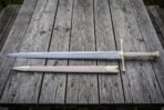 Hand forged fantasy longsword with an engraved double edged blade and brown leather scabbard laid on weathered grey timber decking in soft daylight