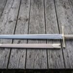 Hand forged fantasy longsword with an engraved double edged blade and brown leather scabbard laid on weathered grey timber decking in soft daylight