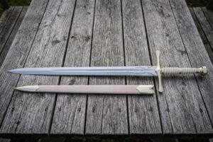 Hand forged fantasy longsword with an engraved double edged blade and brown leather scabbard laid on weathered grey timber decking in soft daylight