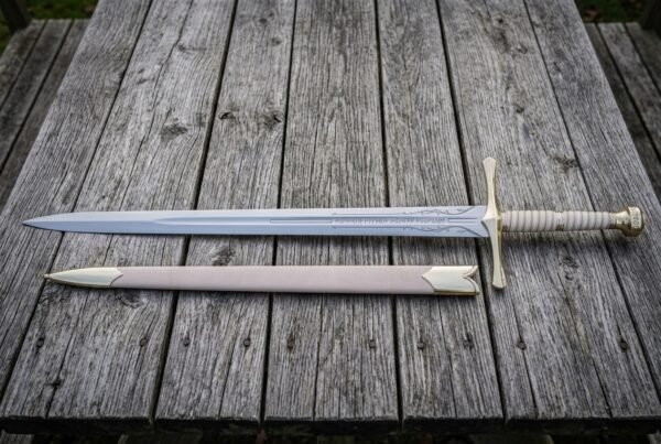 Hand forged fantasy longsword with an engraved double edged blade and brown leather scabbard laid on weathered grey timber decking in soft daylight