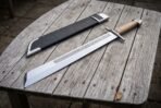 Hand forged grosse messer sword with a broad polished single edged blade and pale hardwood grip lying on a silvered timber table beside a black leather sheath