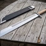 Hand forged grosse messer sword with a broad polished single edged blade and pale hardwood grip lying on a silvered timber table beside a black leather sheath