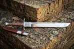 Hand forged grosse messer with a long polished straight backed blade and rosewood grip resting on a lichened stone ledge beside a brown leather sheath