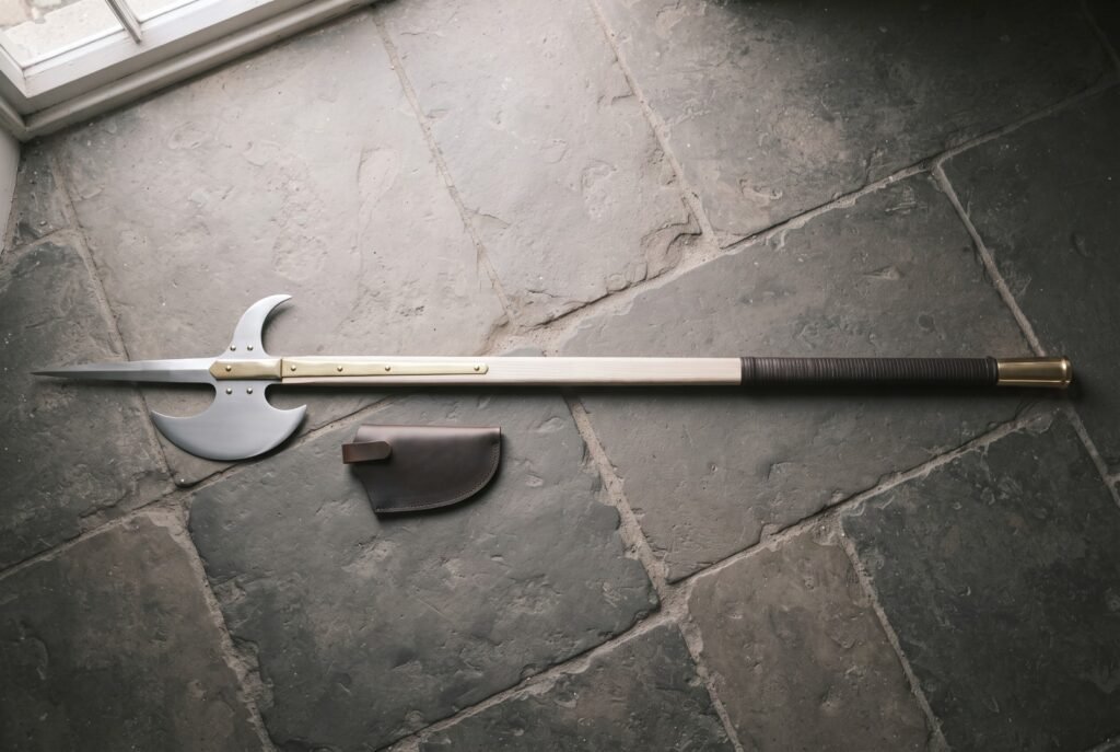 Hand forged halberd with a polished steel spike, crescent blade and rear fluke on an ash haft with brass langets lying on grey flagstones by a leather cover