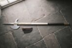 Hand forged halberd with a polished steel spike, crescent blade and rear fluke on an ash haft with brass langets lying on grey flagstones by a leather cover