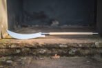 Hand forged high carbon steel glaive polearm with curved blade and pale wooden haft, lying along a stone step by a doorway