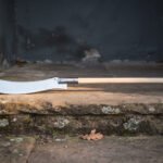 Hand forged high carbon steel glaive polearm with curved blade and pale wooden haft, lying along a stone step by a doorway