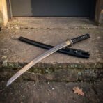 Hand forged high carbon steel katana with black cord wrapped handle and brass tsuba, laid across its black lacquered saya on a stone doorstep