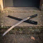 Hand forged high carbon steel katana with black cord wrapped handle and brass tsuba, laid across its black lacquered saya on a stone doorstep