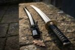 Hand forged high carbon steel katana with black wrapped handle and brass tsuba, close angled view beside its saya on old stonework