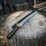 Hand forged high carbon steel katana with brass tsuba and black saya, angled close view of the handle on a tree stump